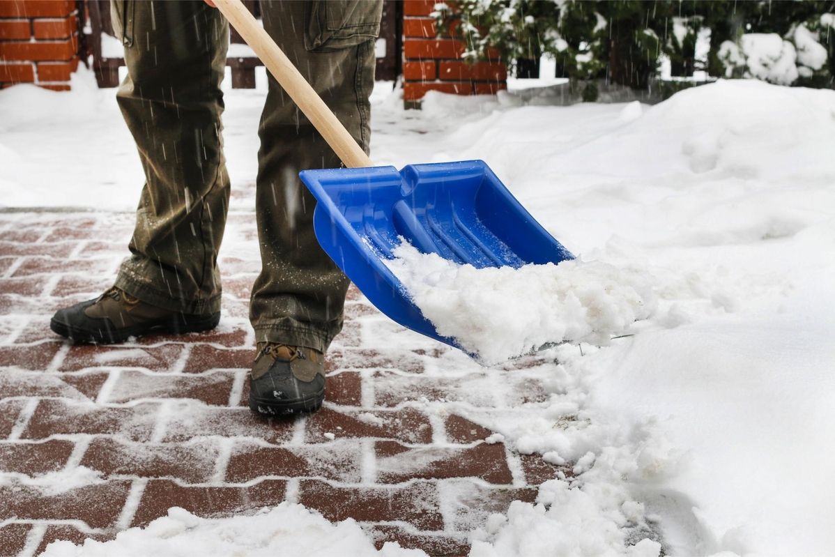 Person schaufelt mit einer blauen Schaufel Schnee von einem gemauerten Weg.